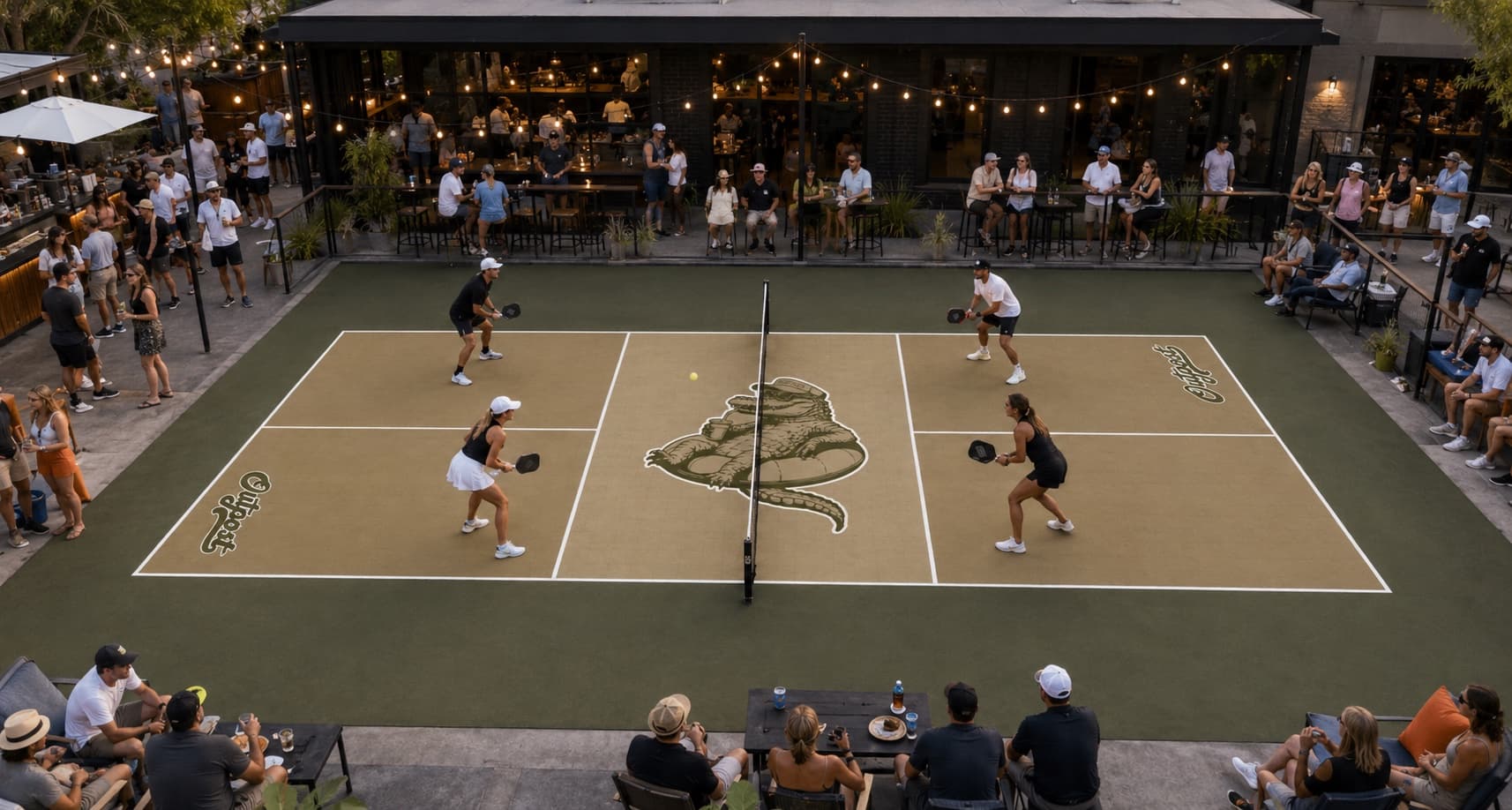 A crowd watching doubles on an Outpost-branded pickleball court at dusk, with the gator logo at centre court and a full patio bar behind it